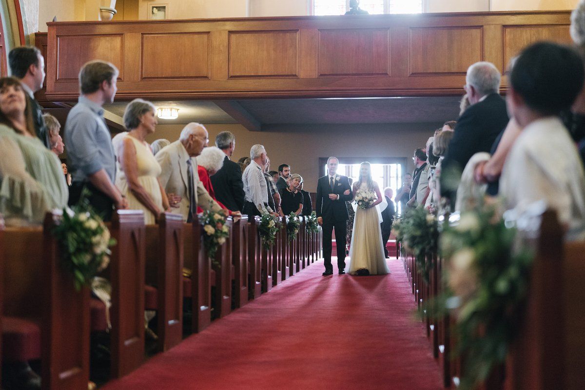 Bride walking down a red-carpeted aisle in a church, accompanied by her father, guests seated on either side.
