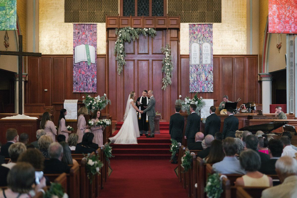 Wedding ceremony in a church: bride and groom exchanging vows, surrounded by guests, altar with floral arch.