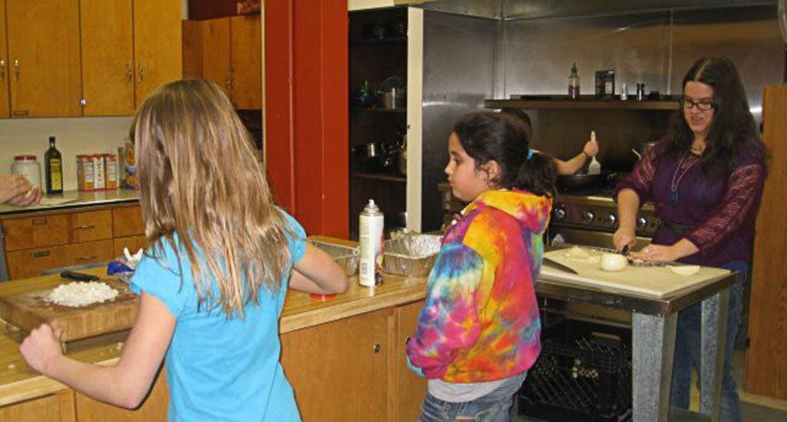 Three children and an adult in a kitchen. One girl chops dough, another watches. The adult cuts onions.