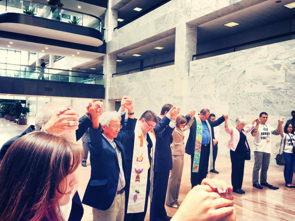 People in a lobby holding hands, raising arms in prayer. Multi-ethnic group, some wearing stoles, natural lighting.