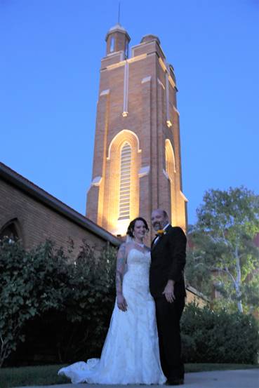 Newlyweds pose in front of a tall brick church tower at dusk; bride in white gown, groom in a tuxedo.