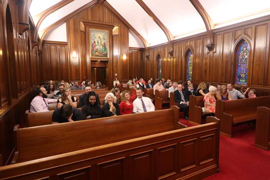 Guests seated in a church for a wedding. Wooden pews, stained glass windows, and an altar with a painting are visible.