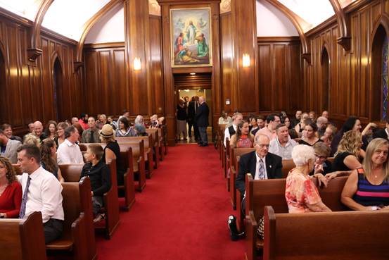 People seated in wooden pews, facing forward in a wood-paneled church. Red carpet aisle.