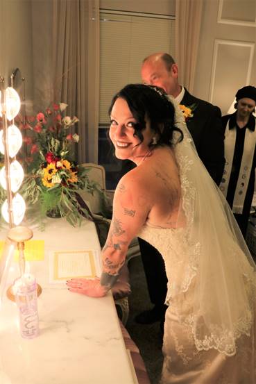 Bride in wedding dress smiles at the camera in a dressing room. A man and a woman stand behind her.