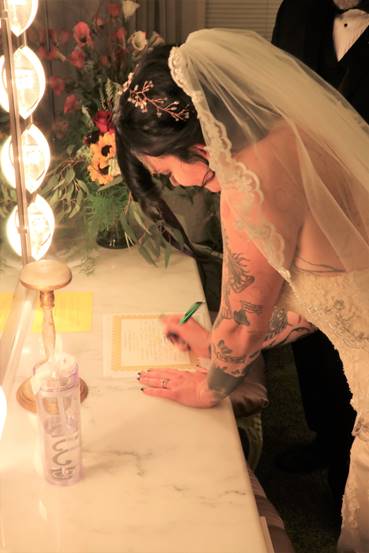 Bride in white gown signing marriage certificate, arm tattoos visible, next to a man in a tuxedo.
