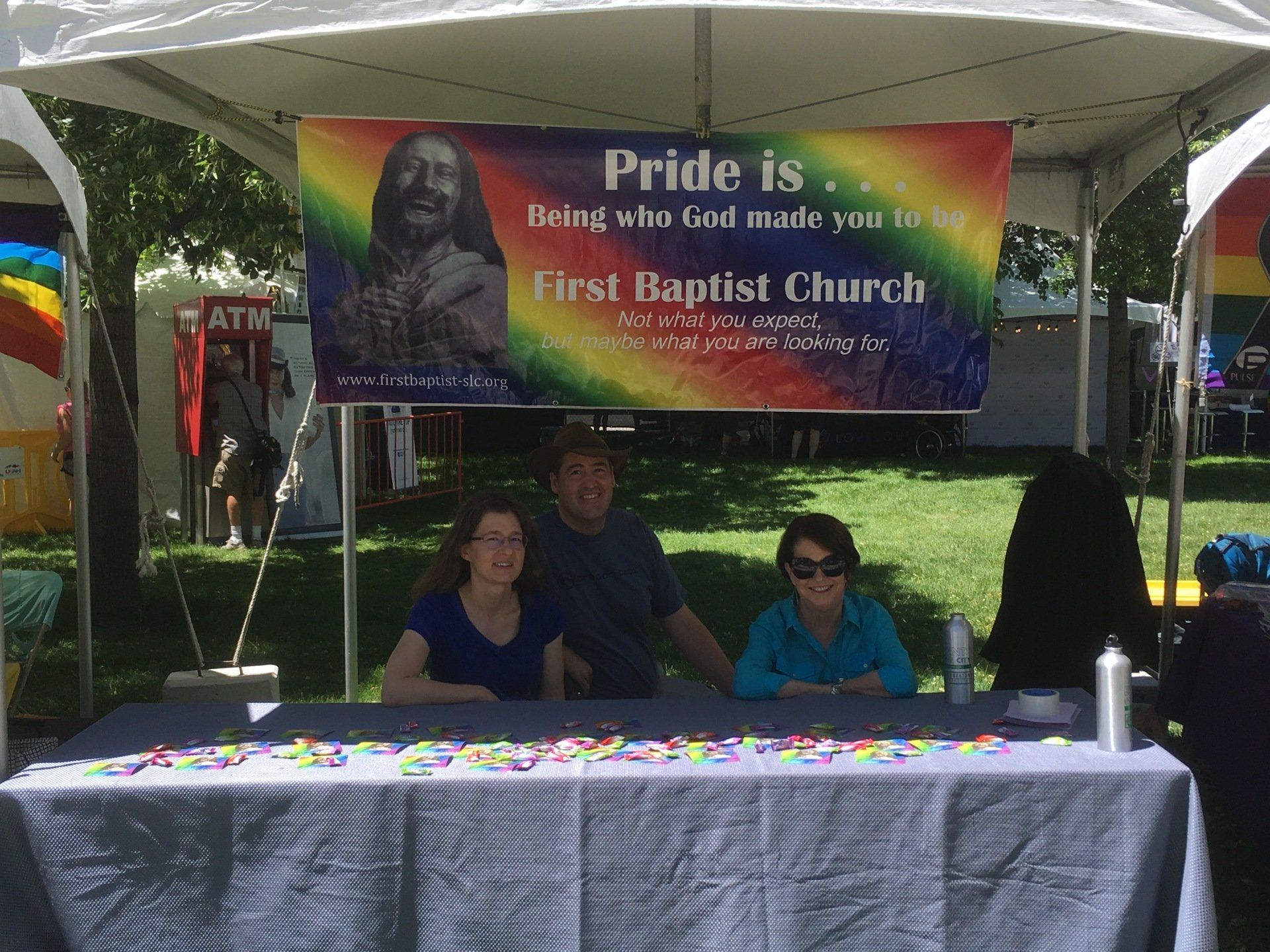 People at a First Baptist Church booth at a Pride event. Rainbow banner: