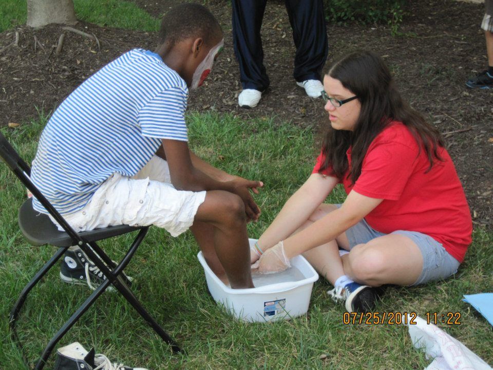 Woman in red shirt washing a Black man's feet in a white basin on grass. Both are outdoors.
