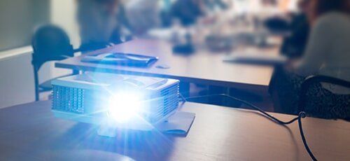 A projector is on a table in a meeting room, shining brightly. People are blurred in the background.