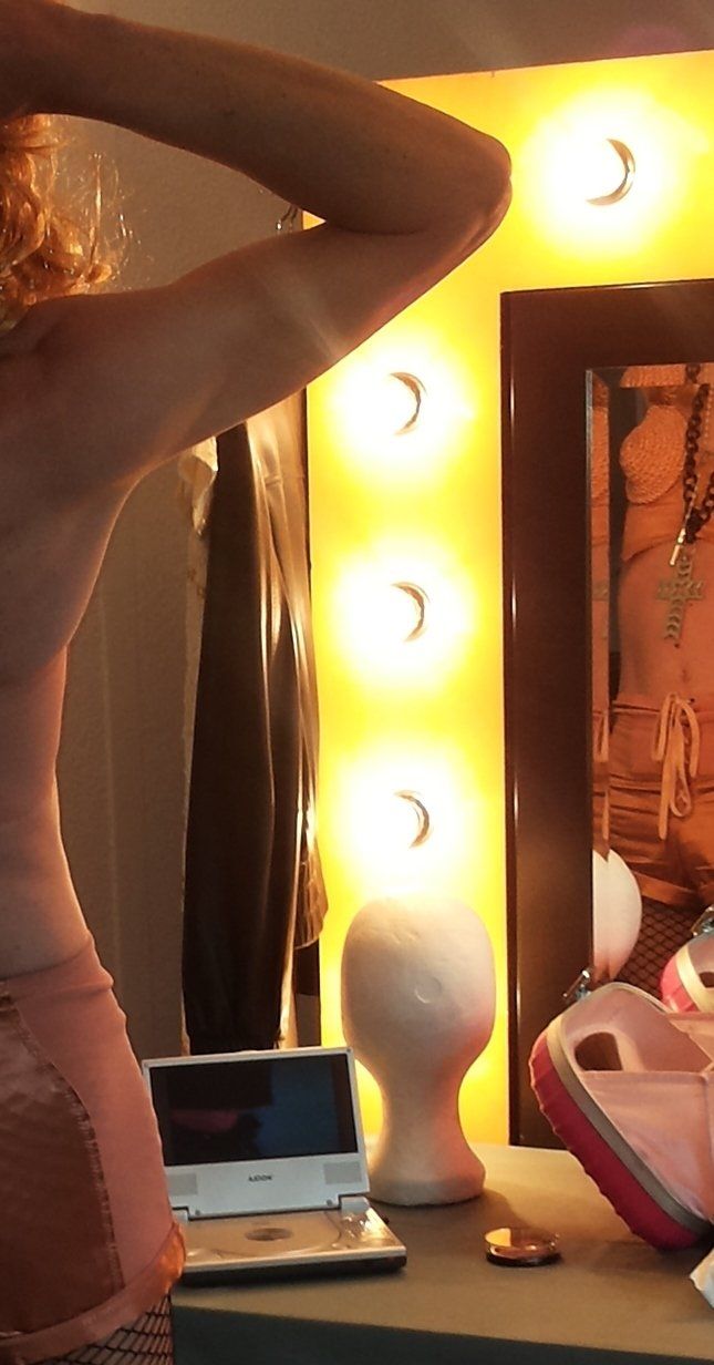 Het Vaarwel van Miss Mighty M. Vrouw in roze jurk staat bij een kapspiegel met verlichting.