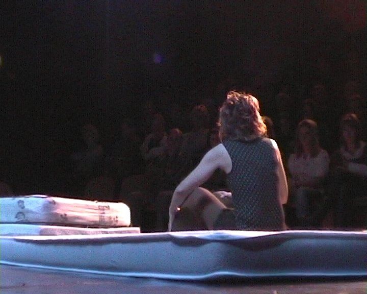 Bobby. Een persoon zit op een matras op een podium, met zijn rug naar de camera. Publiek op de achtergrond.