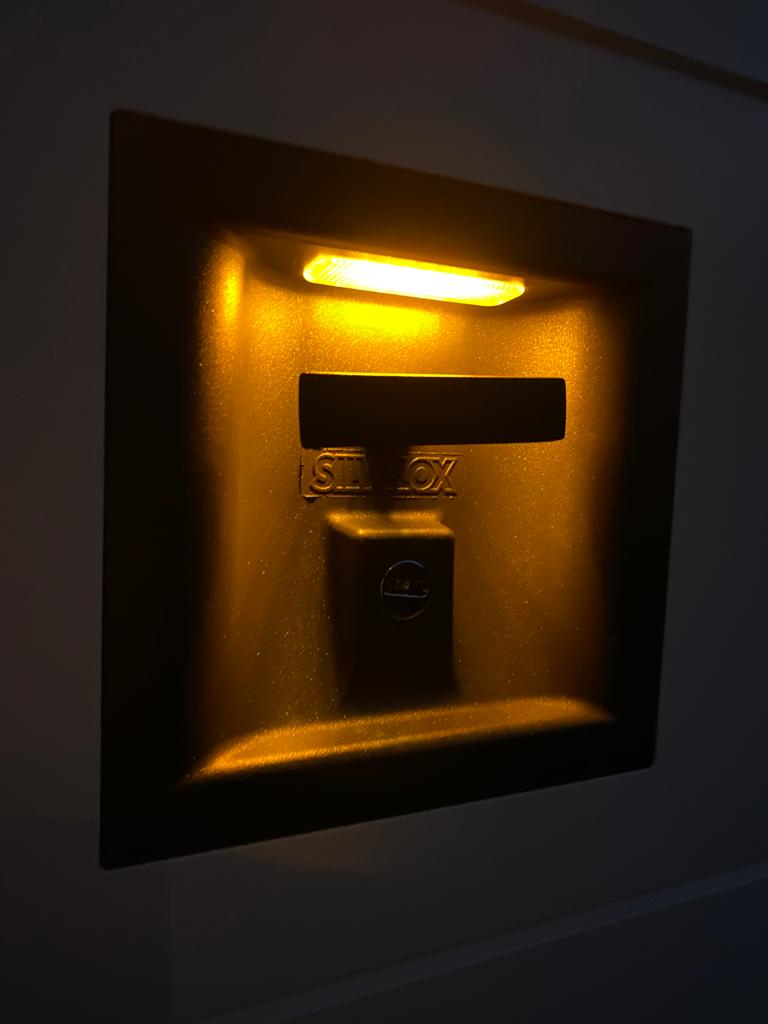 Una lampada da parete a incasso con pareti scure, caratterizzata da una piccola luce orizzontale diffusa posizionata sopra un interruttore centrale.