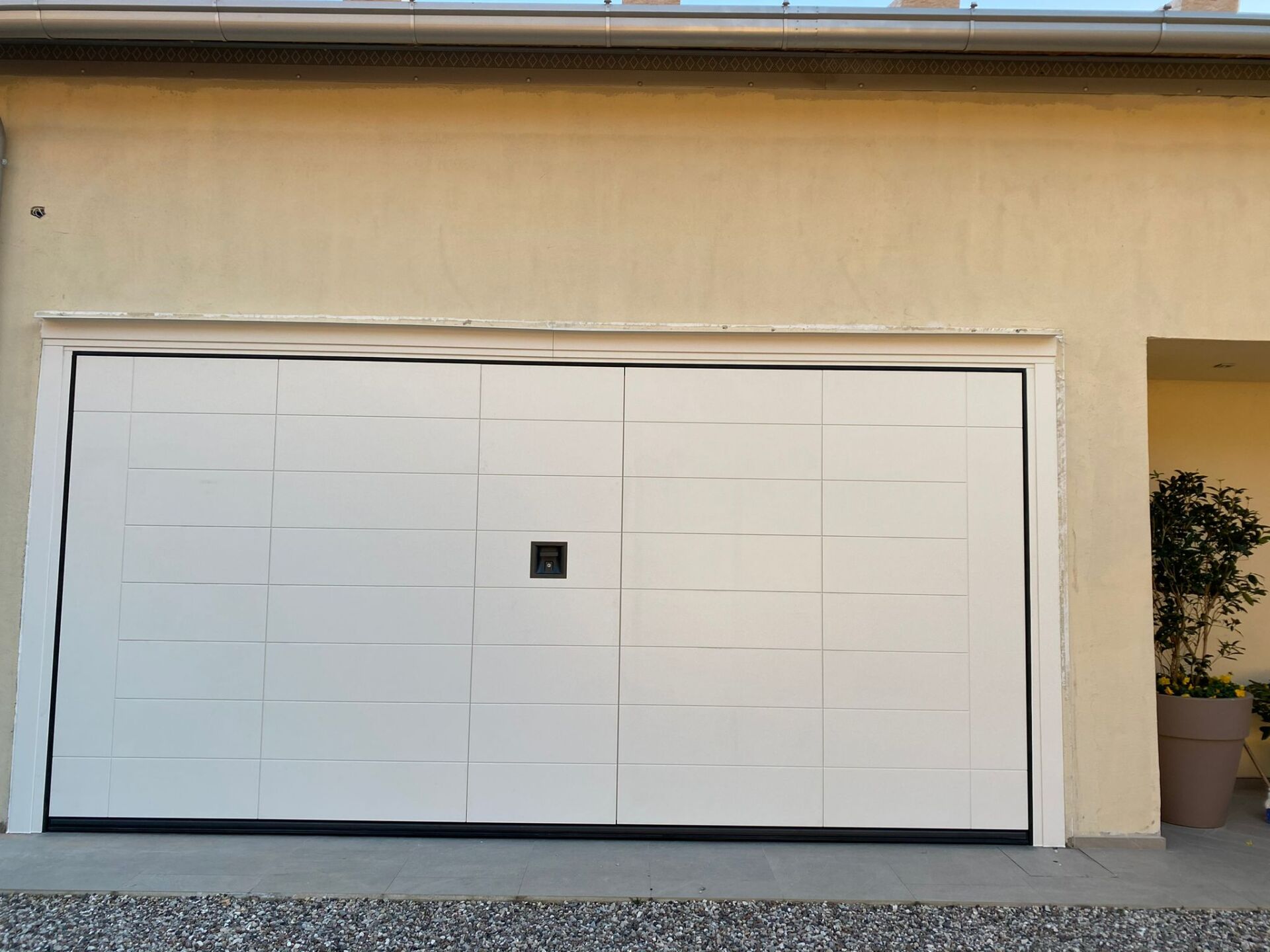 Una moderna porta da garage bianca con motivo a griglia rettangolare e serratura centrale, incastonata in una parete esterna intonacata di beige.