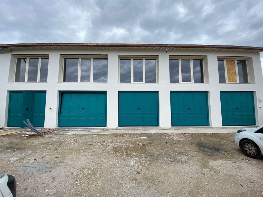 Esterno di un moderno edificio a due piani con cinque portoni da garage color verde acqua e cinque finestre al piano superiore, sullo sfondo di un lotto di ghiaia.