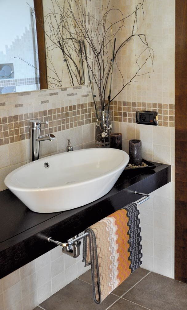 Bagno moderno con lavabo ovale bianco, piano di lavoro scuro e ramoscelli decorativi. Portasciugamani con asciugamani a fantasia.