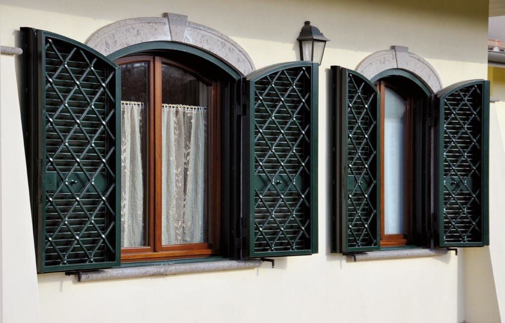 Due finestre con persiane verdi aperte a rombi. Parete beige, dettagli in pietra ad arco, cornici in legno.