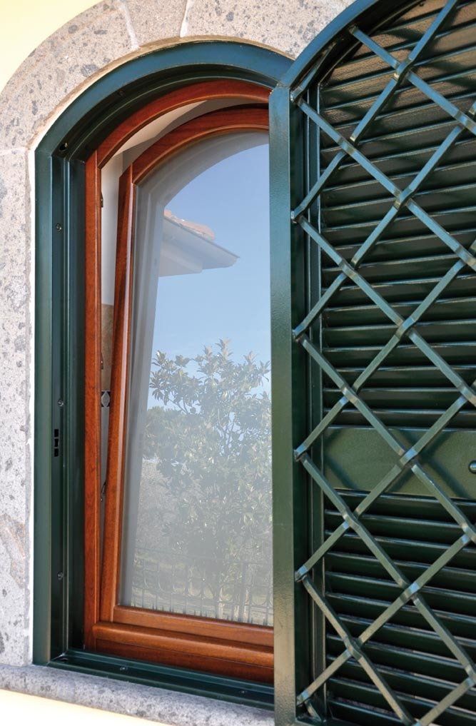 Finestra ad arco con finiture verdi, persiana verde parzialmente aperta. La luce del sole si riflette sul vetro.