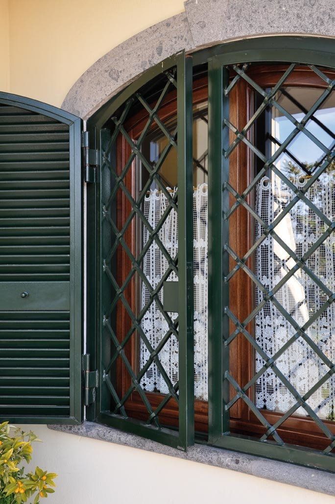 Finestra verde con sbarre di sicurezza a forma di rombo, persiana aperta e tenda bianca.