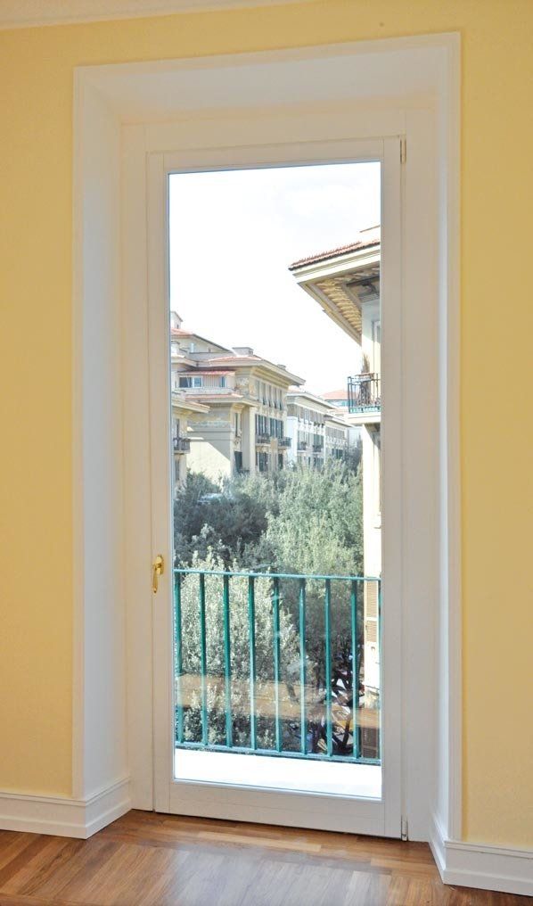 Porta con cornice bianca che si apre su un balcone con ringhiera verde; sono visibili edifici e alberi.