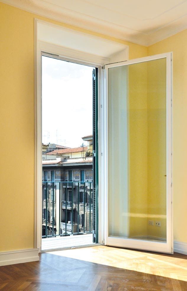 Finestra bianca aperta su un balcone con vista sugli edifici; pareti gialle, pavimento in legno, luce del sole.