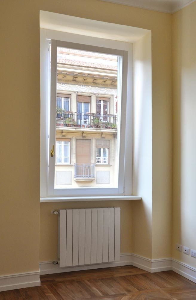 Finestra che si affaccia su un edificio cittadino; cornice bianca, pareti color crema, termosifone, pavimento in legno.
