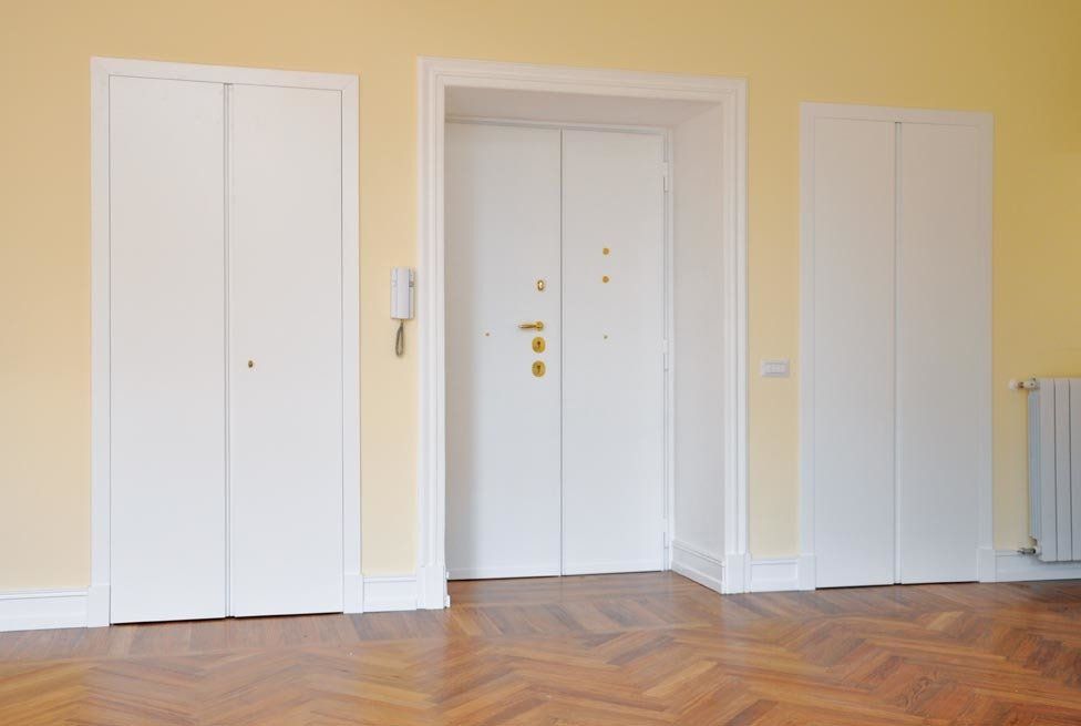 Porta d'ingresso bianca affiancata da due porte abbinate in una stanza con pavimento in parquet e pareti gialle.