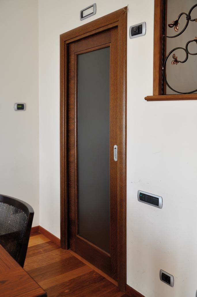 Porta in legno con pannello in vetro smerigliato in una stanza dalle pareti bianche, circondata da interruttori della luce e pavimento in legno.