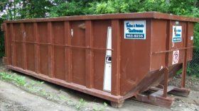 Brown dumpster on the ground, likely for construction debris. Has company logos and a pull-down ramp.