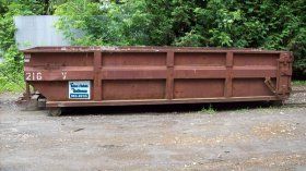 Brown industrial dumpster on a gravel surface near greenery.