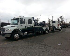 Tow truck towing a white semi-truck cab on a paved lot, overcast day.