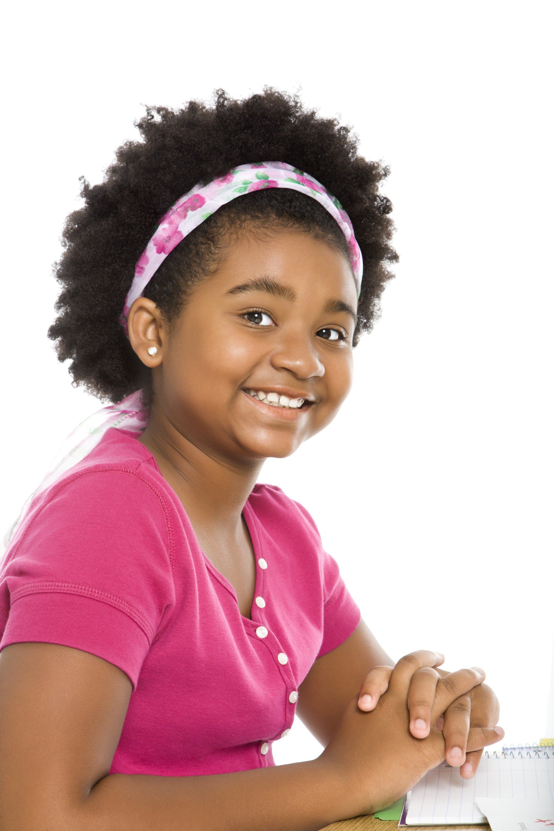 A smiling child with dark curly hair and a patterned headband wears a pink shirt, sitting with hands clasped together.