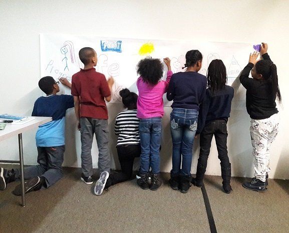Seven children stand and kneel side-by-side against a white wall, drawing on a large paper mural with colorful markers.