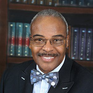 A person wearing glasses and a polka-dot bow tie, smiling in front of a bookshelf.
