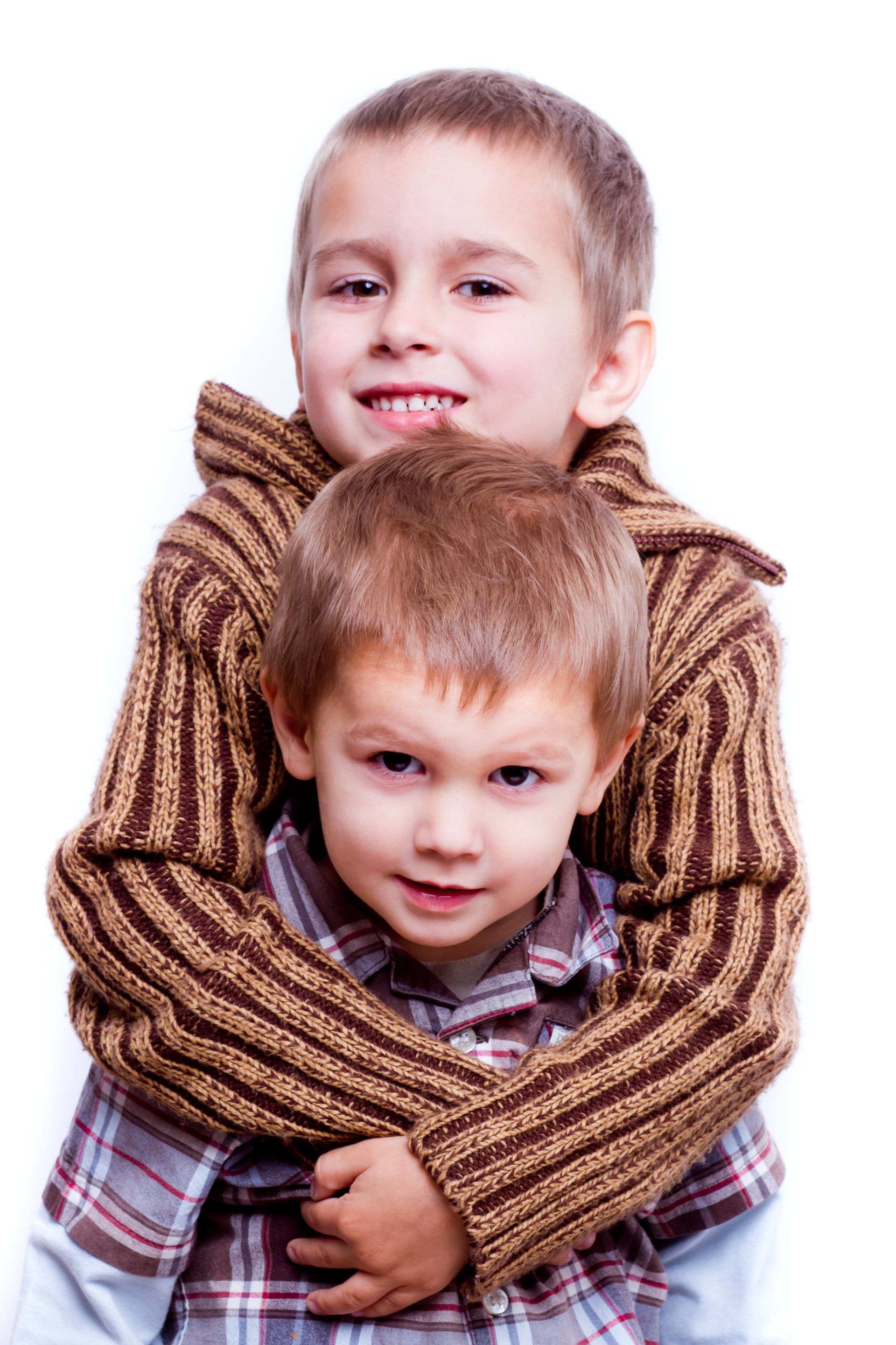 Two children smiling, with one hugging the other from behind, both wearing textured sweaters against a white background.