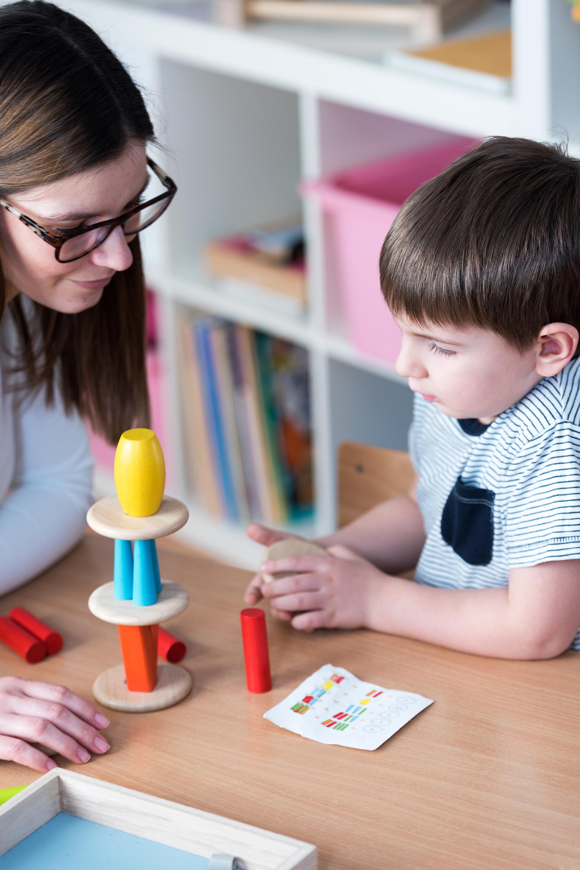A teacher and student sit at a table building a colorful wooden tower with blocks while following a pattern card.