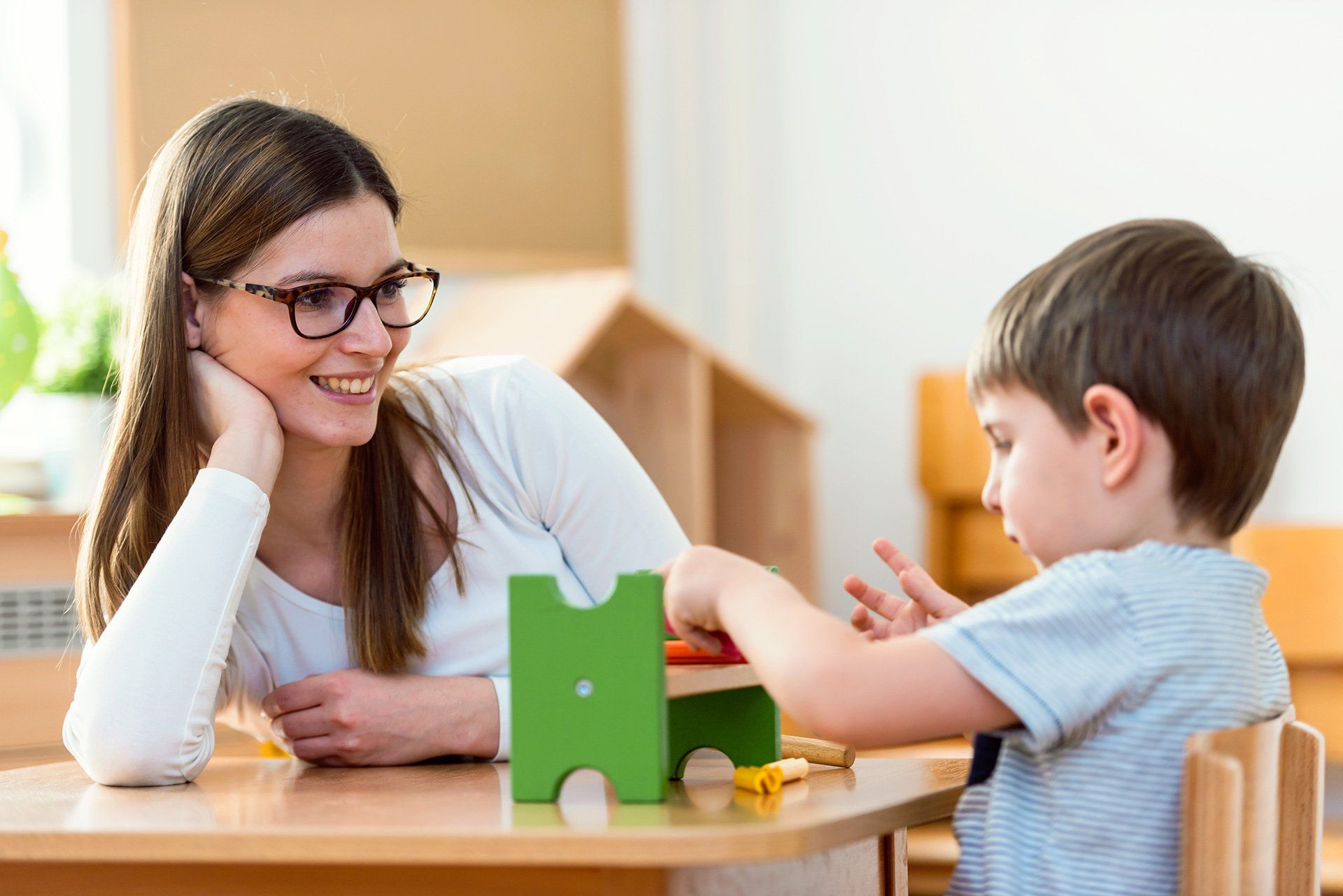 A person with glasses watches a child playing with a green toy at a wooden table in a room.