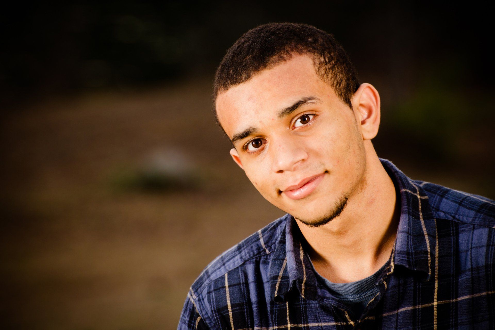 A person wearing a blue plaid shirt looks toward the camera with a soft smile against a blurry, dark outdoor background.