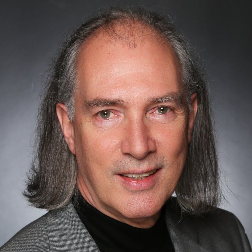 Headshot of a person with shoulder-length gray hair wearing a black shirt and gray blazer against a gray background.