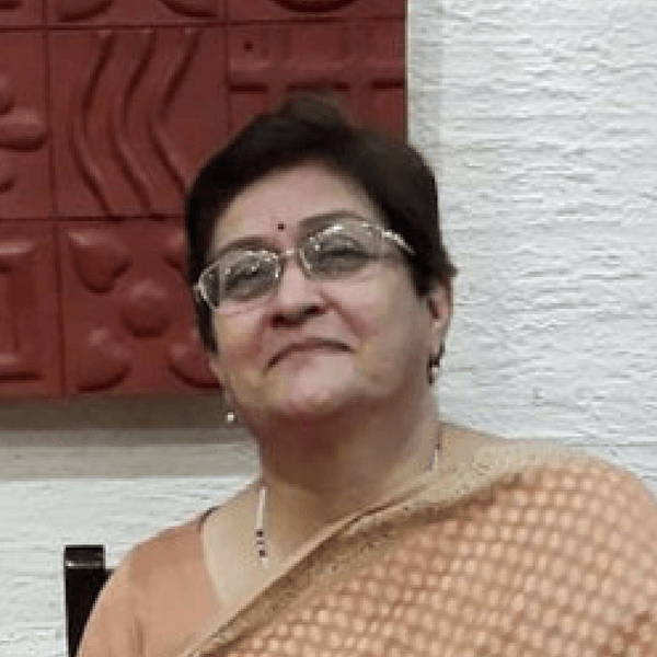 A person wearing glasses and a light-colored sari smiling, positioned in front of a red decorative wall.