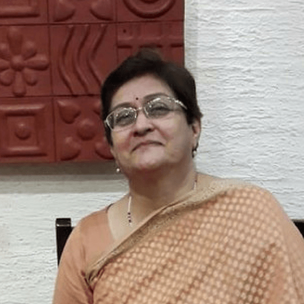 A person with short dark hair and glasses wears a light peach sari, posed against a textured white and red wall.