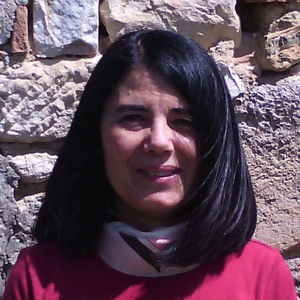 A smiling person with dark hair and a red top, posed in front of a rustic stone wall.