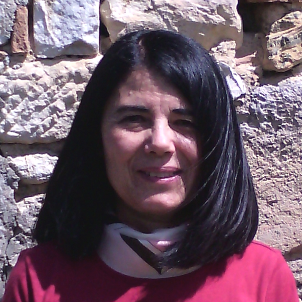 A person with shoulder-length dark hair wearing a red shirt and a neck brace, posing against a textured stone wall.