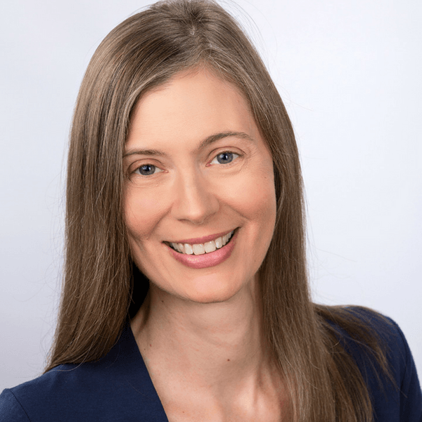 A professional headshot of a smiling person with long brown hair wearing a dark blue blazer against a white background.