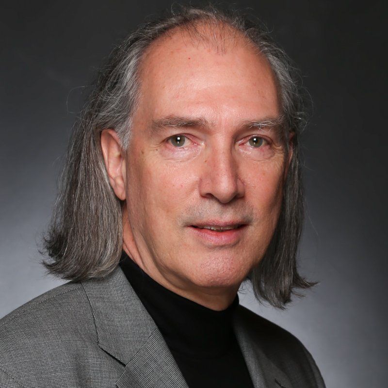 Headshot of a man with shoulder-length gray hair, wearing a black turtleneck and gray blazer against a dark background.