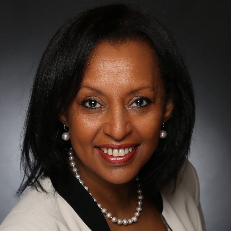 A professional headshot of a smiling person wearing a cream-colored blazer and a pearl necklace against a dark background.