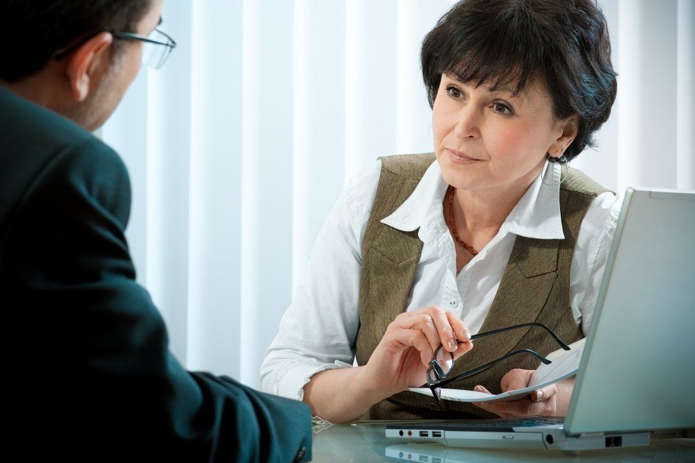 Two professionals sitting at a desk during a meeting, with one holding glasses and reviewing documents near a laptop.