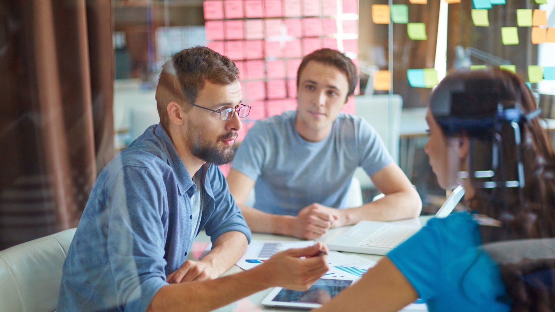 Three people collaborate around a table in an office, reviewing documents and a tablet near a wall of sticky notes.