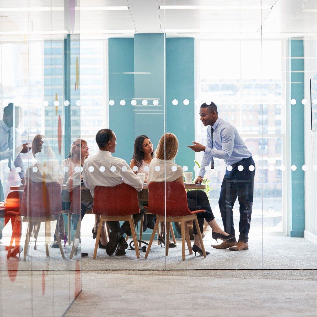 Colleagues in business attire meet in a bright office with glass walls, as one person stands to speak to the seated group.