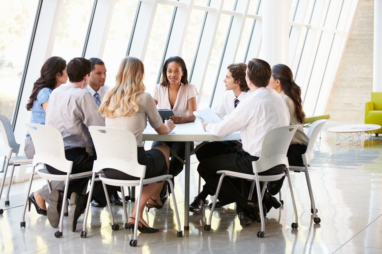 A group of professionals sits around a table in a brightly lit, modern office with floor-to-ceiling windows, in a meeting.