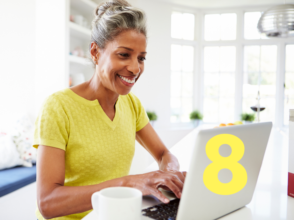 A person in a yellow shirt smiles while typing on a laptop in a bright kitchen.