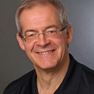 A smiling person with gray hair and glasses wearing a black shirt, set against a gray background.